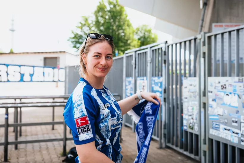 Porträt einer lächelnden Frau in blau-weißer Sportbekleidung mit Schal vor Stadionkulisse