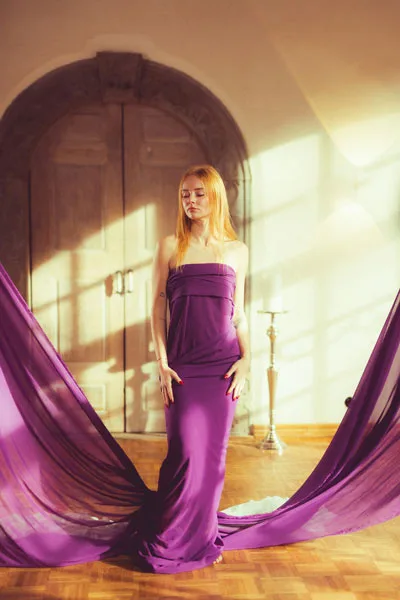 Portrait einer blonden Frau in einem lila trägerlosen Kleid vor einem hellen Rundbogen.