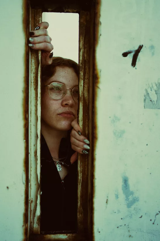 Portrait einer Frau mit Brille, die durch einen schmalen Schlitz in rostigem Metall blickt