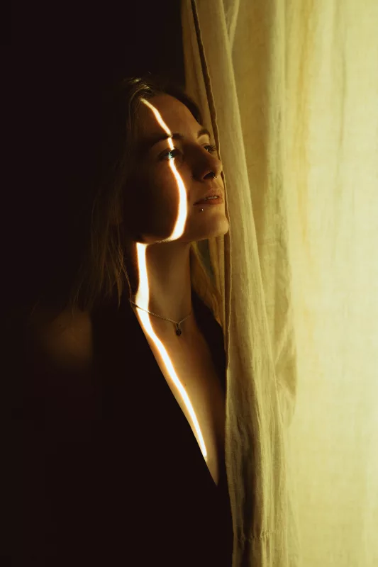 Portrait einer Frau, deren Gesicht durch einen schmalen Lichtstreifen hinter einem Vorhang beleuchtet wird.