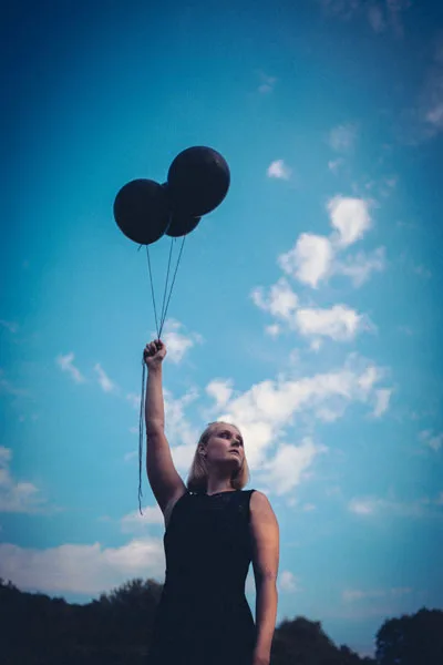 Portrait einer Frau, die drei schwarze Luftballons gegen einen blauen Himmel hochhält.