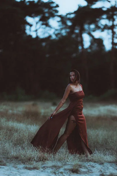 Portrait einer Frau in einem fließenden, dunkelroten Kleid auf einer Wiese bei Dämmerung.