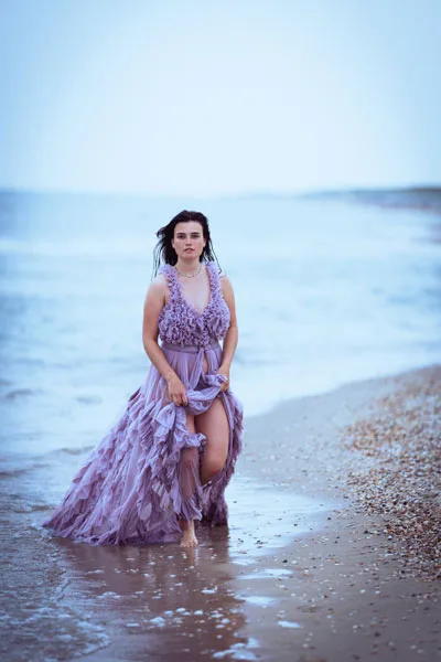 Portrait einer Frau in einem fließenden lila Kleid im seichten Meer am Strand.