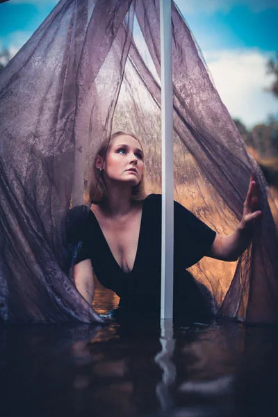 Portrait einer Frau hinter fließendem, violettem Stoff mit Spiegelung im Wasser.