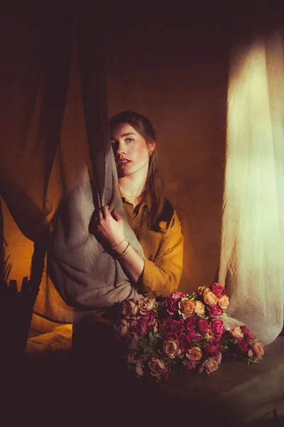 Portrait einer Frau hinter einem hellen Stoffvorhang mit rosa Rosen im Vordergrund.