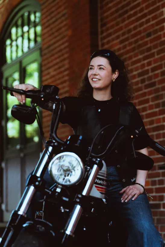 Portrait einer lächelnden Frau mit Lederweste auf einem Motorrad vor einer Backsteinwand