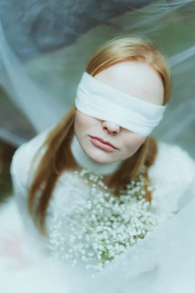 Portrait einer Frau mit rötlichem Haar und weißer Augenbinde, die weiße Blumen hält