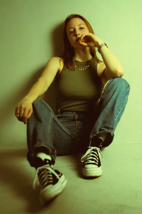 Portrait einer jungen Frau in einem olivgrünen Top und Jeans, sitzend auf dem Boden.