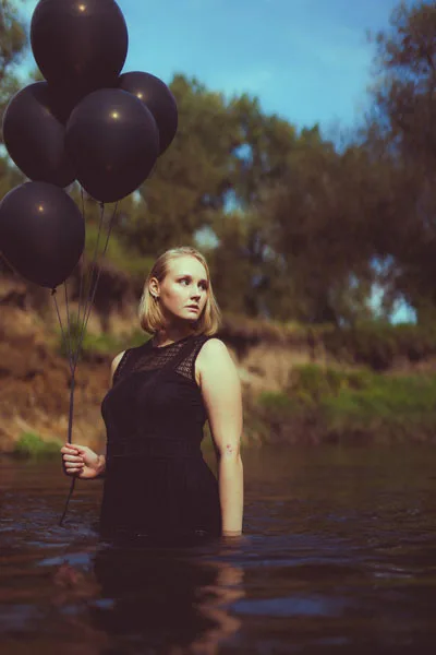 Portrait einer Frau in schwarzem Kleid mit schwarzen Luftballons im Wasser.