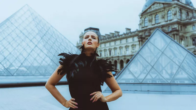 Portrait einer Frau im schwarzen Kleid mit Federbesatz vor der Glaspyramide des Louvre in Paris.