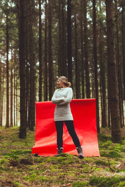 Portrait einer Frau in weißem Pullover vor einem großen roten Hintergrund im Wald.