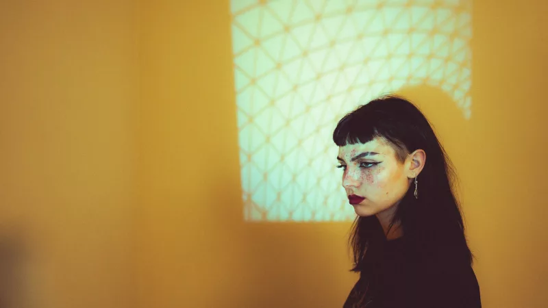 Portrait einer jungen Frau mit dunklem Haar und Sommersprossen vor einer Wand mit geometrischer Lichtprojektion.
