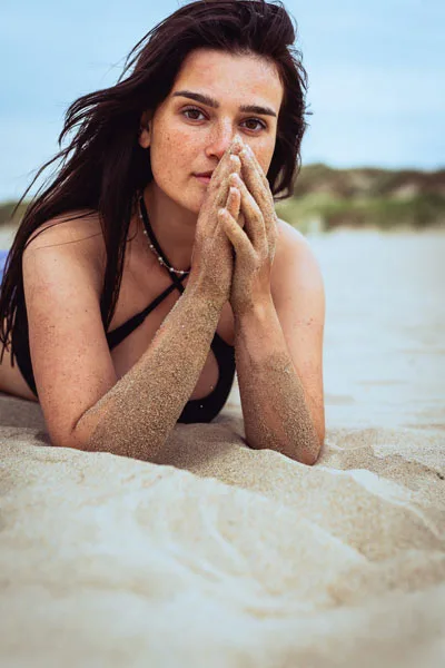 Portrait einer jungen Frau mit dunklen Haaren und Sommersprossen, die im Sand liegt.