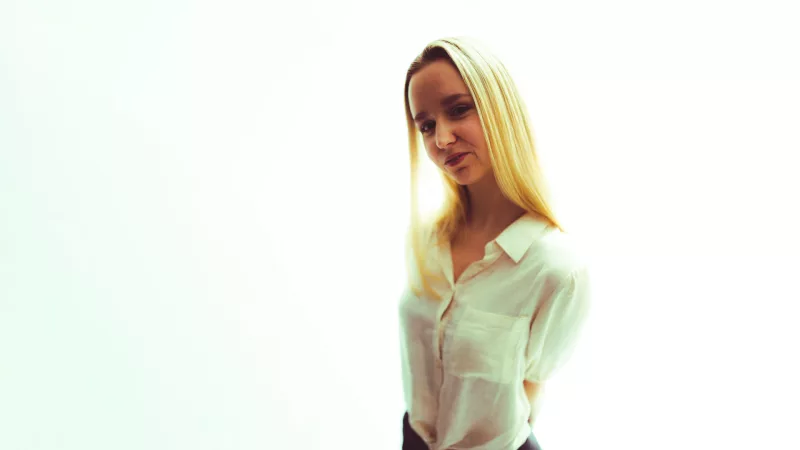 Portrait einer jungen Frau mit langen blonden Haaren und weißer Bluse vor hellem Hintergrund.