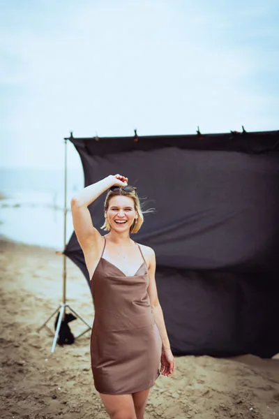 Portrait einer lächelnden Frau in einem braunen Satinkleid vor schwarzem Hintergrund am Strand.