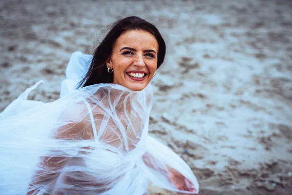 Portrait einer lächelnden Frau mit weißem, fließendem Schleier am Strand.