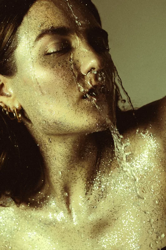 Portrait einer Person mit glitzernder Haut und herabfließendem Wasser im Gesicht.