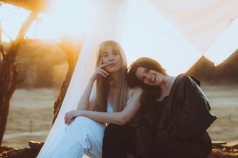 Portrait zweier Frauen im warmen Licht des Sonnenuntergangs unter weißem Stoff, eine lehnt ihren Kopf lächelnd an die Schulter der anderen.