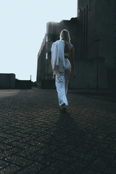 Rückansicht einer Frau in weißem Outfit vor massiver, dunkler Architektur.