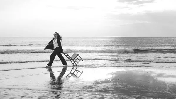 Schwarz-weiß-Aufnahme einer Frau, die einen kleinen Holzstuhl am Strand entlangzieht.