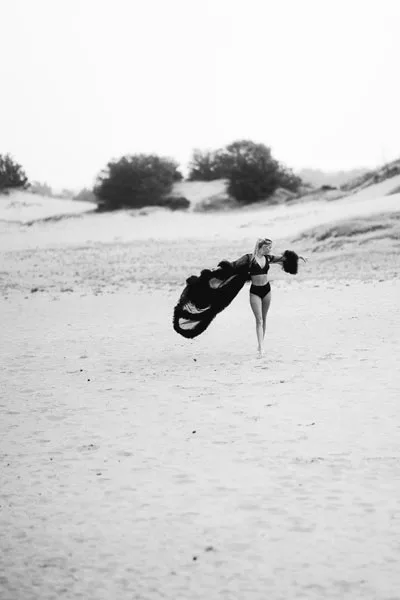 Schwarz-weiß-Aufnahme einer Frau am Strand mit einem im Wind wehenden dunklen Stoff.
