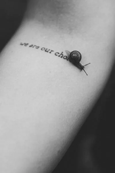 Schwarz-weiß Detailaufnahme eines Tattoos mit dem Schriftzug „we are our choices“ und einer kleinen Schnecke auf der Haut.