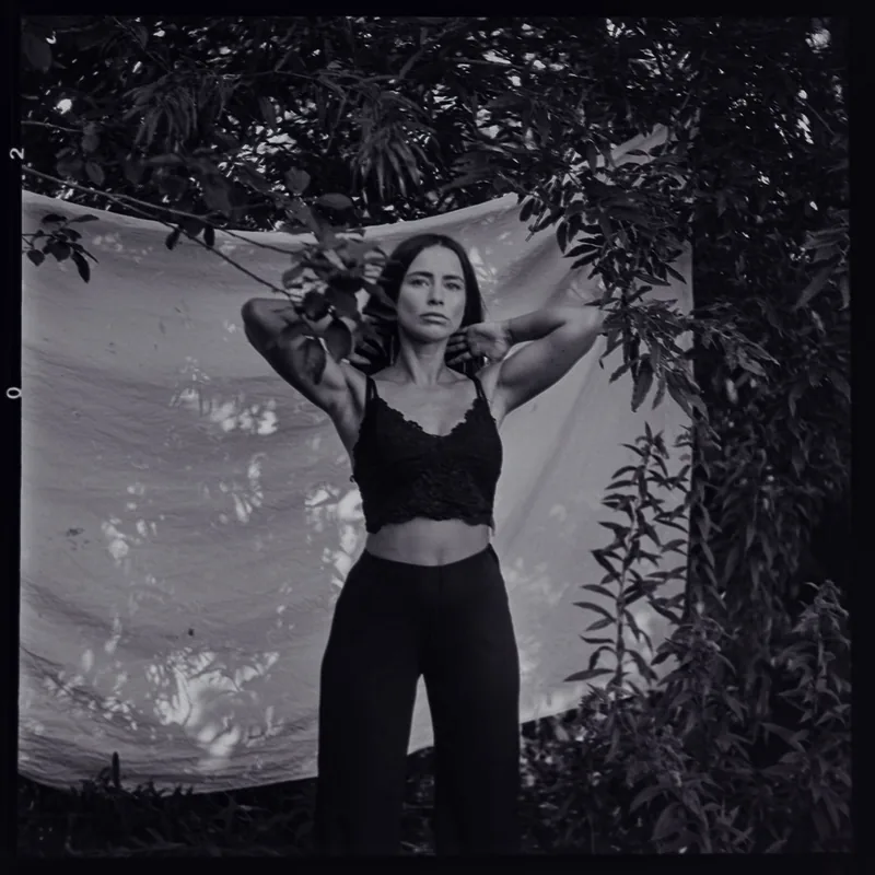 Schwarz-weiß Portrait einer Frau in dunklem Crop-Top vor einem hellen Stoffhintergrund und Blättern.