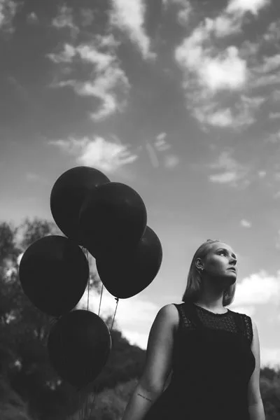 Schwarz-weiß Portrait einer Frau mit dunklen Luftballons vor einem bewölkten Himmel.