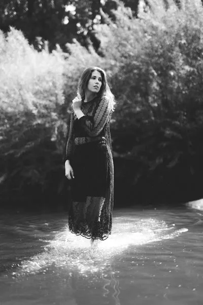 Schwarz-weiß-Portrait einer Frau in einem dunklen Spitzenkleid, die im seichten Wasser vor dichtem Blattwerk steht.
