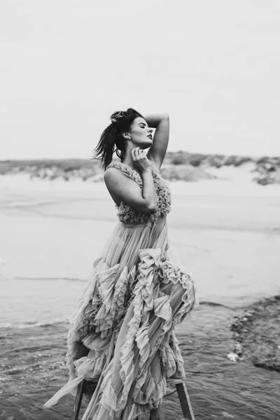 Schwarz-Weiß-Portrait einer Frau in einem fließenden, rüschigen Kleid am Strand.