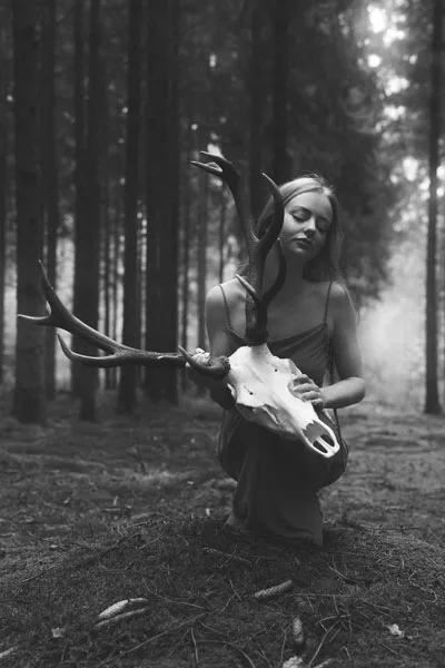 Schwarz-weiß Portrait einer Frau mit einem Hirschschädel in einem dunklen Wald