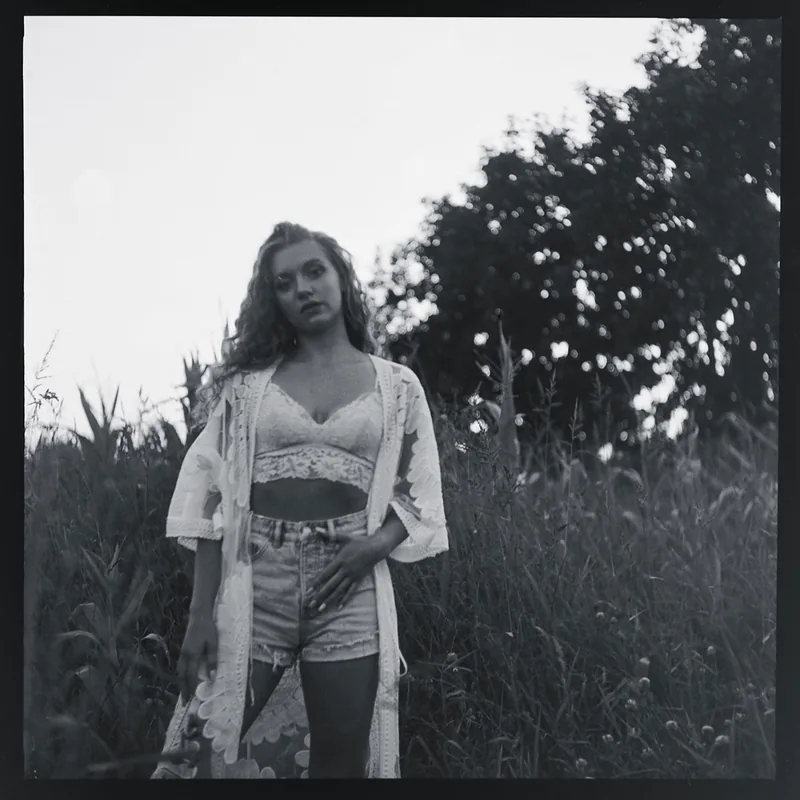 Schwarz-weiß Portrait einer Frau mit lockigem Haar in einem Feld, gekleidet in Spitzenoberteil und Jeansshorts.