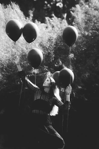 Schwarz-Weiß-Portrait einer Frau mit mehreren dunklen Luftballons vor einem natürlichen Hintergrund.