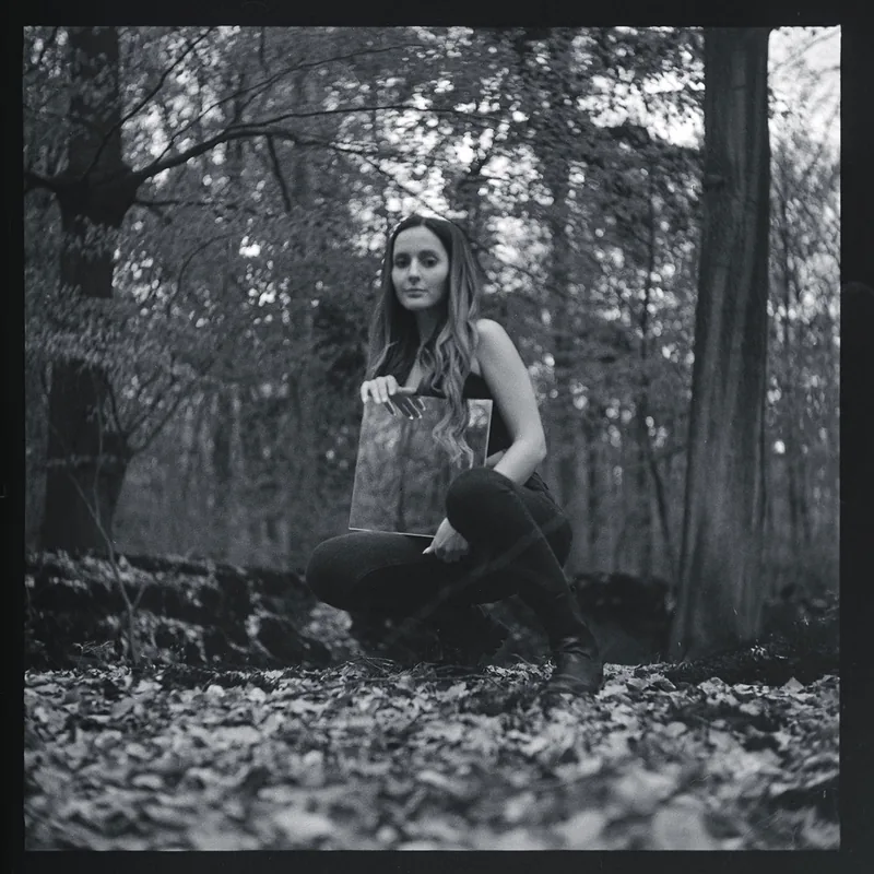 Schwarz-weiß-Portrait einer Frau, die in einem Wald ein Bild hält.