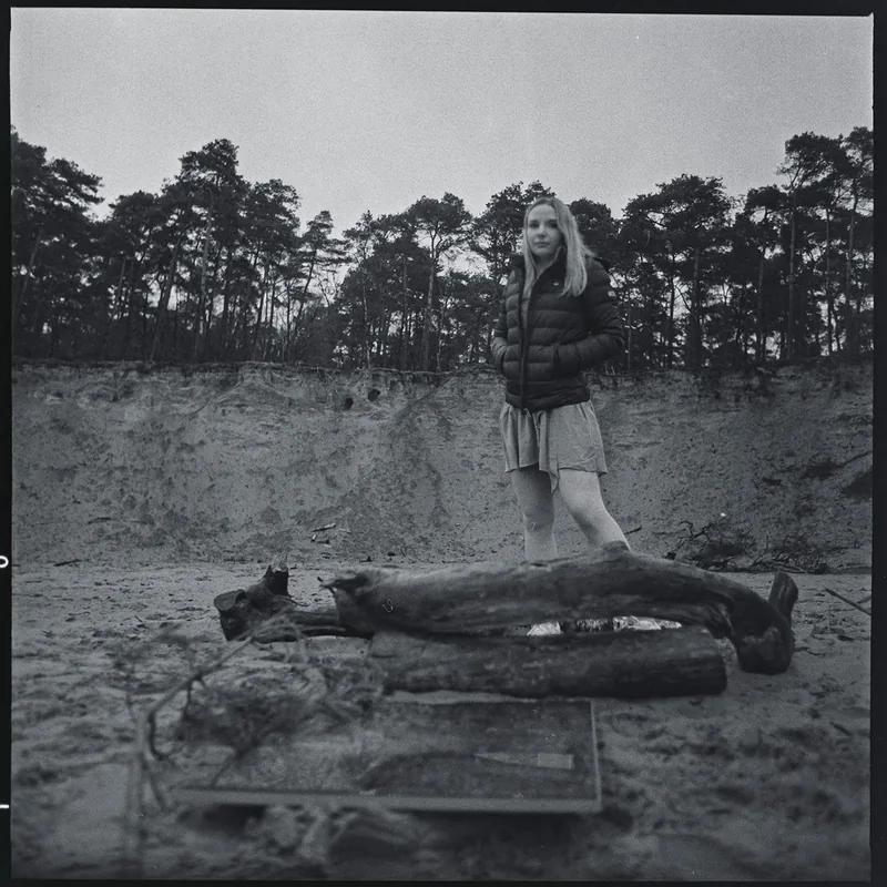 Schwarz-weiß-Portrait einer jungen Frau in einer sandigen Waldlandschaft neben Baumstämmen.