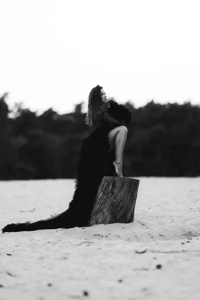 Schwarz-weiß-Portrait einer Person in einem langen dunklen Kleid auf Treibholz am Strand.