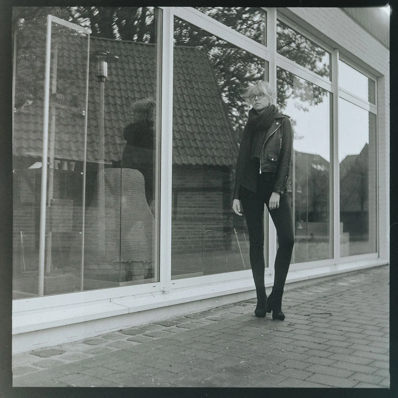 Schwarz-weiß-Portrait einer Person in Lederjacke und Brille vor einer spiegelnden Glasfront.