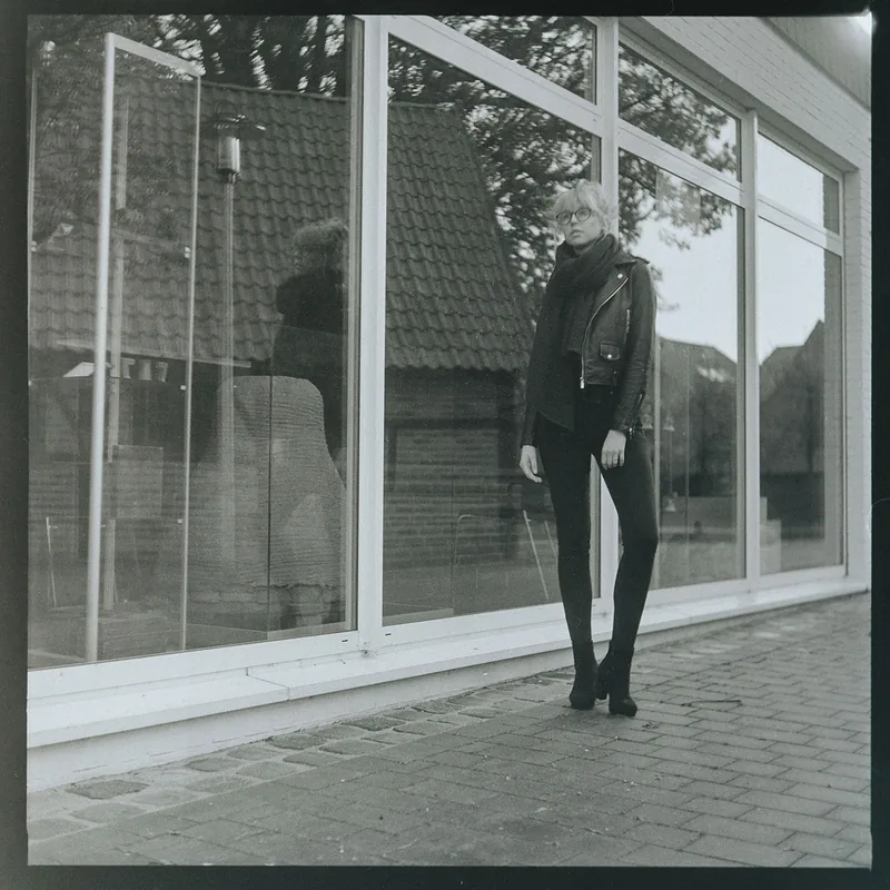 Schwarz-weiß-Portrait einer Person in Lederjacke und Brille vor einer spiegelnden Glasfront.