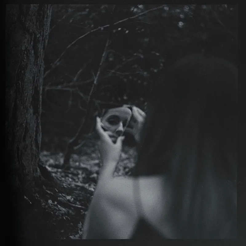 Schwarz-Weiß-Portrait einer Person im Wald, die ihr Gesicht in einem kleinen Spiegel betrachtet.
