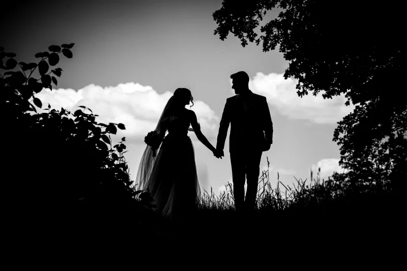 Schwarz-weiß Silhouette eines Brautpaares, das Hand in Hand vor einem bewölkten Himmel steht.