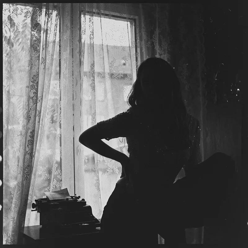 Schwarz-weiß Silhouette einer Frau vor einem Fenster mit Spitzenvorhängen und einer Schreibmaschine.