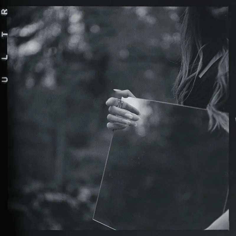Schwarz-weißes Analogportrait einer Hand, die eine reflektierende Glasscheibe vor unscharfem Laub hält.