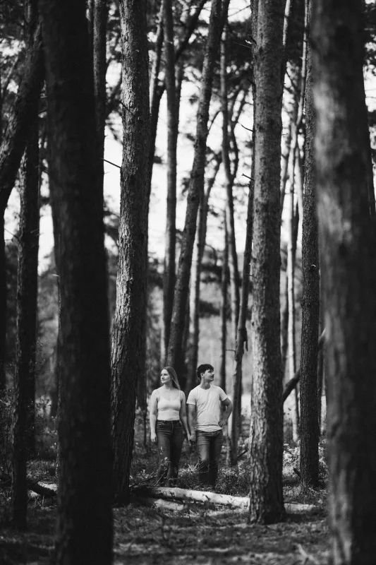 Schwarz-weißes Paarshooting zwischen hohen Bäumen im Wald.