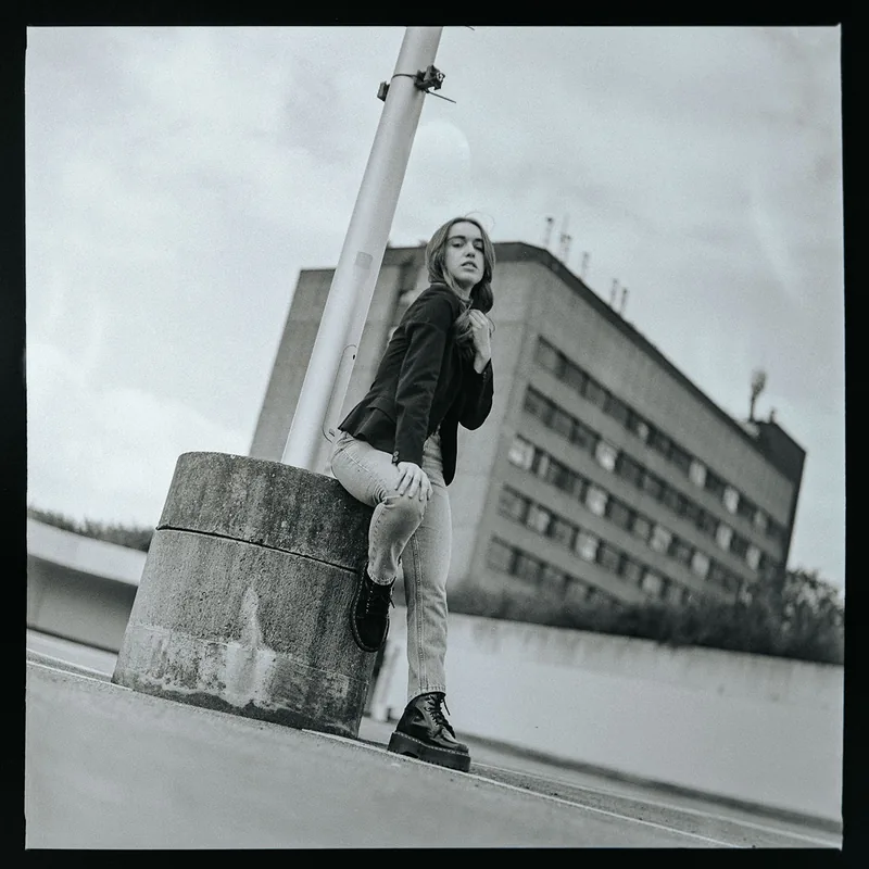 Schwarz-weißes Portrait einer Frau, die an einem Betonsockel lehnt, vor einem urbanen Gebäude.