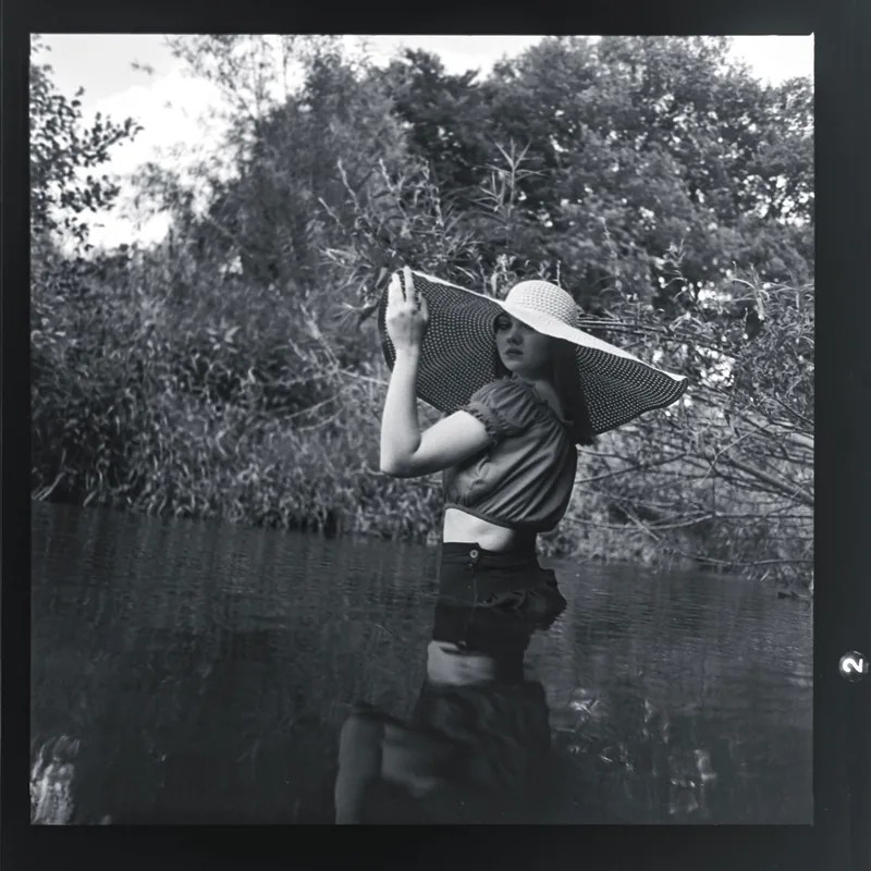 Schwarz-weißes Portrait einer Frau mit großem Sonnenhut im Wasser zwischen Schilf und Bäumen.