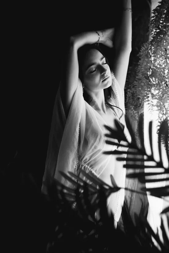 Schwarz-weiß Boudoirportrait einer Frau mit erhobenen Armen hinter Farnwedeln