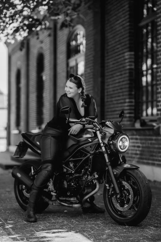 Schwarzweiß-Portrait einer lächelnden Frau auf einem Motorrad vor einer Backsteinwand
