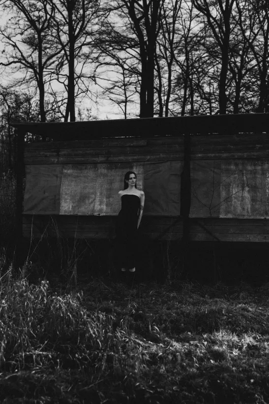 Schwarzweiß-Portrait einer Frau in einem dunklen, trägerlosen Kleid vor einer rustikalen Holzwand im Wald