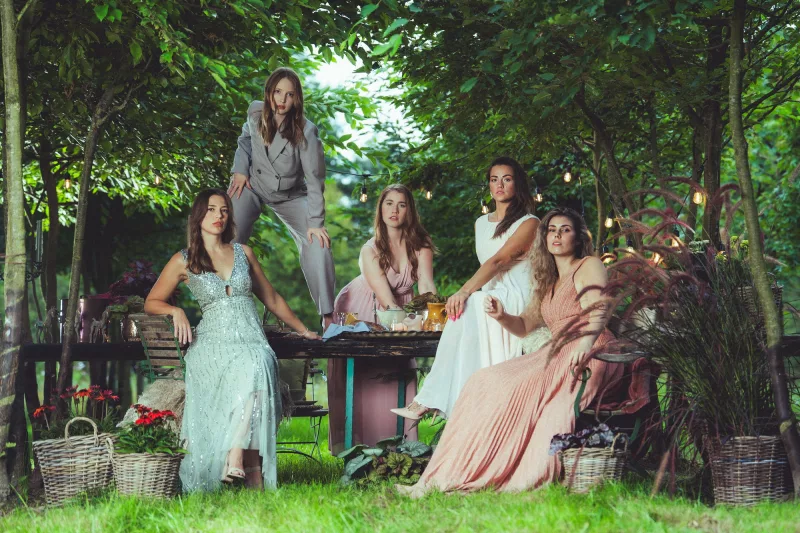 Sechs Frauen in eleganter Kleidung posieren für ein Gruppenfoto in einer grünen Gartenkulisse.