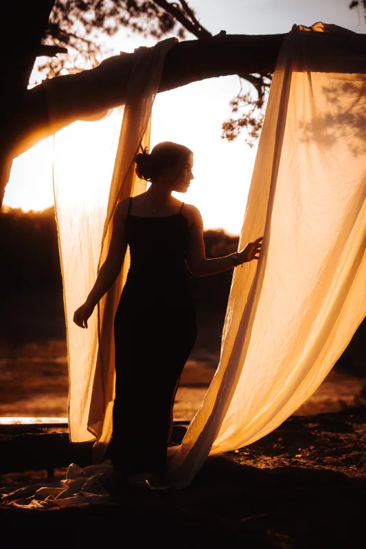 Silhouette einer Frau zwischen lichtdurchlässigen Stoffbahnen im warmen Licht des Sonnenuntergangs.